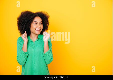 Happy happy hispanic o brasiliana capelli ricci donna, gesturing con pugni, gioire nel successo, realizzazione, vincere, guarda a lato al copy-space per pubblicizzare, sfondo giallo isolato Foto Stock