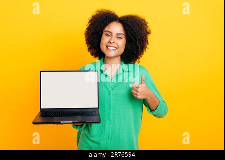 Donna curly brasiliana o afro-americana positiva, in camicia verde, tenendo in mano un portatile aperto con schermo bianco bianco mock up, sorridere alla fotocamera, mostra il gesto pollice su, sfondo giallo isolato Foto Stock