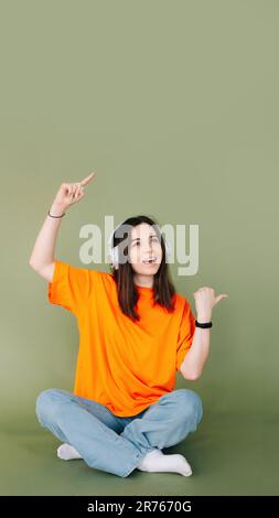 Ritratto di una donna gioiosa che indossa le cuffie, esprimendo entusiasmo e approvazione puntando entrambi i pollici verso uno spazio vuoto, isolato su un Foto Stock