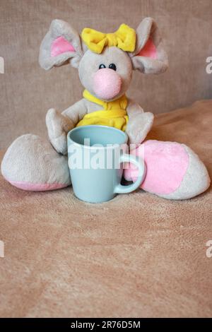 Simpatico giocattolo con tisana seduto sul divano. Giocattolo morbido per animali. Concetto di Buongiorno. Background infantile. Camera per bambini. Divertente giocattolo morbido del mouse. Foto Stock