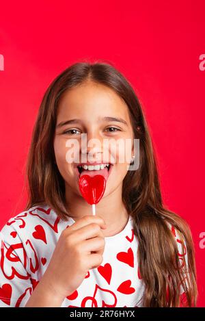 Ritratto di positiva carina bambina in capelli lunghi e abiti casual leccando lollipop rosso e guardando la macchina fotografica mentre si è in piedi contro sfondo rosso Foto Stock