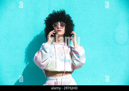 Seria giovane donna ispanica con acconciatura afro parlare sul telefono cellulare e toccare i capelli con la mano mentre si è in piedi e guardando la macchina fotografica Foto Stock