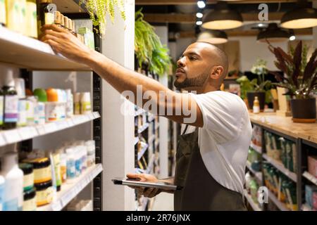 Vista laterale di concentrato giovane afro-americano venditore maschile in grembiule prendendo il prodotto da scaffale mentre in piedi con tablet durante il processo di lavoro in cismo Foto Stock