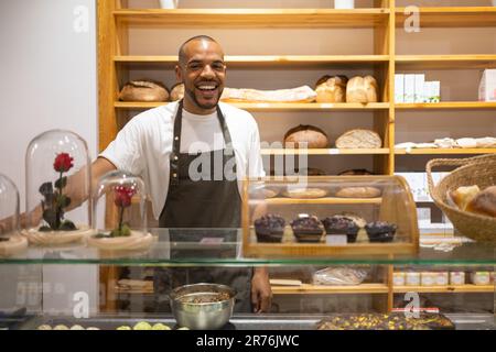 Allegro giovane afro americano venditore maschile in grembiule sorridente e guardando la macchina fotografica mentre si trova dietro il bancone contro gli scaffali Foto Stock