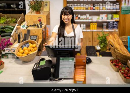 Felice giovane venditore femminile asiatico in grembiule sorridente e utilizzando il computer mentre si trova al banco con vari prodotti durante il lavoro in supermercato Foto Stock