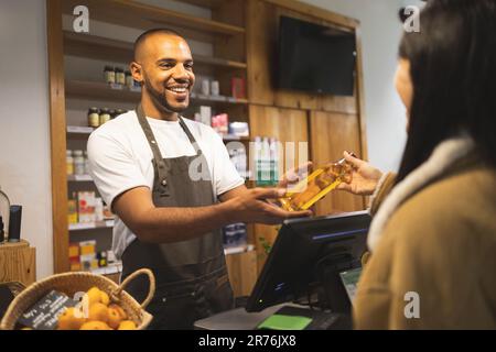 Positivo giovane afro-americano venditore maschile in grembiule sorridente e dando bottiglia di olio al cliente mentre si trova al banco durante il lavoro Foto Stock