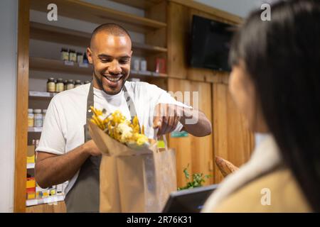 Positivo giovane afro-americano venditore maschile in grembiule sorridente e dando sacchetto di carta con prodotti per anonima cliente femminile in moderno supermercato Foto Stock