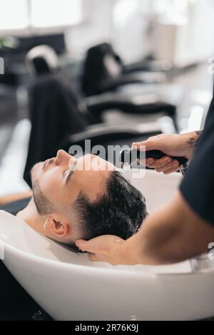 Giovane barbiere anonimo corto lavare i capelli dell'uomo su sfondo sfocato nel barbiere Foto Stock