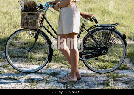 Vista laterale di una femmina tagliata e irriconoscibile che si trova in piedi sul manubrio della bicicletta con cesto di vimini pieno di punte di grano di luce diurna Foto Stock