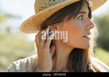 Ritratto di giovane donna sorridente in cappello di paglia che tocca la parte posteriore della testa con le dita e che guarda lontano da sfondo sfocato alla luce del sole Foto Stock