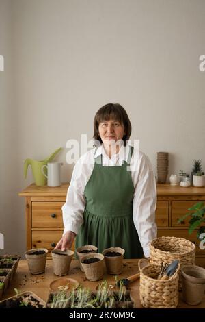 Positiva donna di mezza età in grembiule agricolo sorridendo e guardando la macchina fotografica mentre in piedi vicino al tavolo di legno con pianta in vaso di legno e tazze a casa Foto Stock