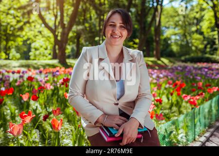 Donna d'affari caucasica felice guardando la macchina fotografica rilassarsi nel parco primaverile. Insegnante che ha una pausa all'aperto rilassarsi nel campus universitario in possesso di libri. Foto Stock