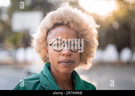 Ritratto di seria afro-americana femmina con capelli ricci e occhiali guardando la macchina fotografica mentre si è in piedi sulla strada con sfondo sfocato Foto Stock