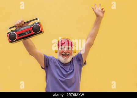 Uomo anziano allegro con fascia rossa e cuffie che tengono il lettore di cassette con un'espressione del volto entusiasta mentre si trova su sfondo giallo Foto Stock