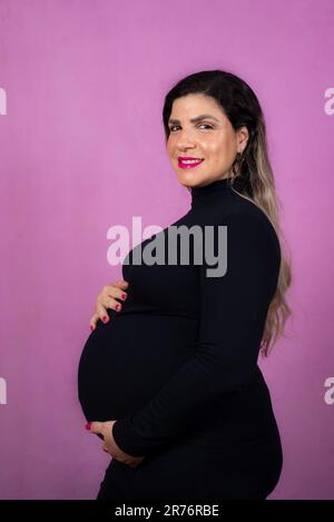 Una donna felice incinta che indossa una parte superiore nera a maniche lunghe si erge su un vivace sfondo viola Foto Stock