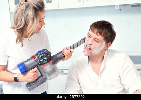 Giovane coppia di famiglia divertirsi in cucina casa. La moglie pulisce il volto del marito, che è coperto di farina, con l'aspirapolvere. L'uomo gli coprì il volto di farina. Foto divertente. Foto Stock