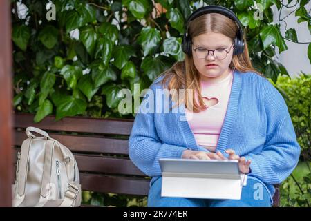 Teenager biondo concentrato in cuffie ascoltare podcast controlla i social network utilizzando il parco notebook Foto Stock