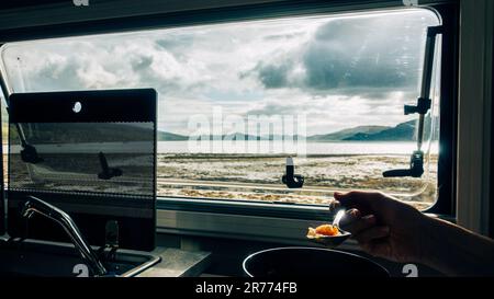 Persona che mangia la cena in un campervan che guarda fuori attraverso la finestra sopra un lago soleggiato nelle Highlands scozzesi su un campo selvaggio in Argyll. Scozia Foto Stock