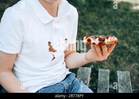 Macchie sporche sui vestiti. Un ragazzo che mangia un dolce ricoperto di cioccolato. all'aperto Foto Stock