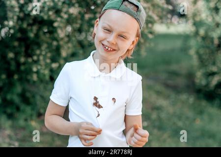 Ritratto faccia sorridente un bambino con macchie di cioccolato sporche su vestiti bianchi. concetto di colorazione della vita quotidiana. all'aperto. Foto Stock