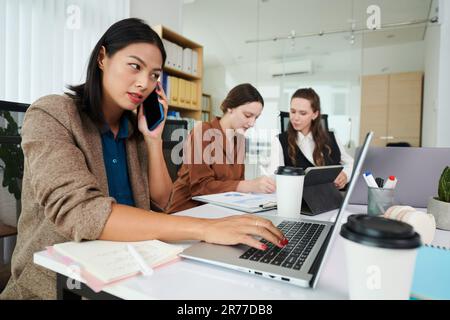 Giovane donna d'affari che parla al telefono cellulare e digita sul computer portatile con i suoi colleghi che lavorano in background Foto Stock