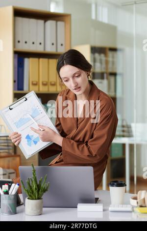 Immagine verticale di una giovane donna d'affari che indica un documento con grafici e ne discute con i colleghi durante una riunione online sul laptop Foto Stock