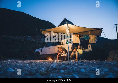 Uomo e donna con cani campeggio di fronte a un veicolo fuoristrada 4x4 con tenda sul tetto di notte e illuminazione romantica sulla spiaggia Foto Stock