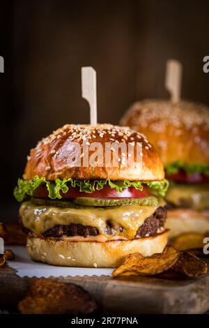 Gustoso cheeseburger con lattuga, formaggio cheddar, pomodoro e sottaceti. Panino hamburger con semi di sesamo. Atmosfera rustica. Delizioso pasto fast food Foto Stock