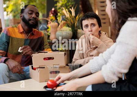Giovane coppia diversa degustazione di verdure fresche di casa prima di acquistare mentre si siede a tavola con il venditore del mercato contadino locale, amici multirazziali uomo e donna in visita fiera biologica, selettiva focus Foto Stock