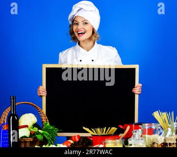 Cuoco femminile sorridente con lavagna bianca. Preparazione del cibo. Menu sano. Cuocete in uniforme da chef con cartellone vuoto. Copia spazio per il menu pubblicizza in Foto Stock