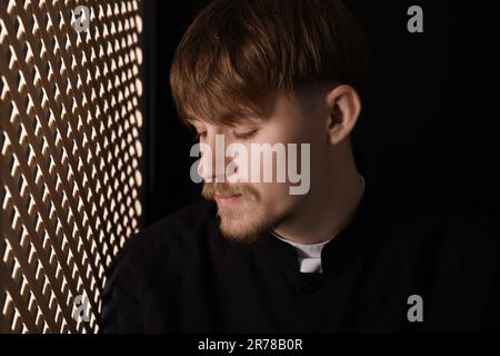 Sacerdote cattolico vicino alla finestra di legno in cabina confessionale Foto Stock