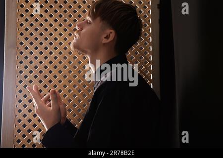 Sacerdote cattolico che prega vicino alla finestra di legno in cabina confessionale Foto Stock