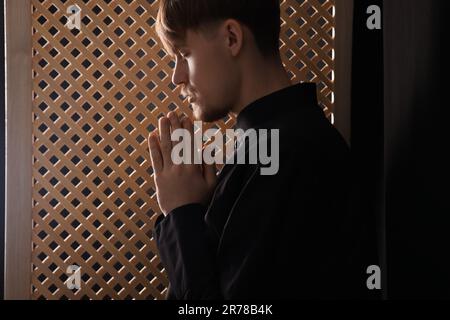 Sacerdote cattolico che prega vicino alla finestra di legno in cabina confessionale Foto Stock