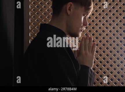 Sacerdote cattolico che prega vicino alla finestra di legno in cabina confessionale Foto Stock