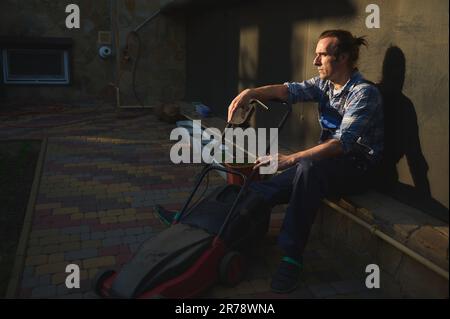 Ritratto di un uomo da giardinaggio stanco sovraccaricato con rasaerba elettrico, rilassante dopo una dura giornata di lavoro nel cortile Foto Stock