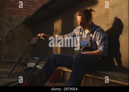 Ritratto di un giardiniere lavorato in tute blu con un rasaerba in giardino al tramonto dopo una dura giornata di lavoro Foto Stock