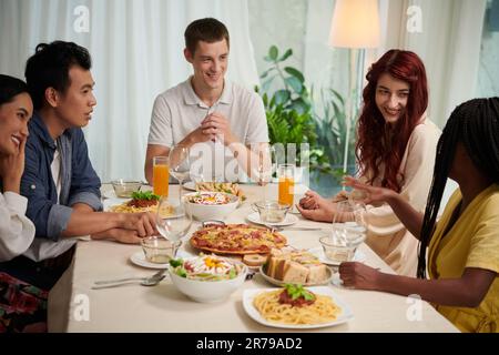 Gruppo di giovani interculturali in casualwear intelligente chiacchierando al tavolo servito e gustando la cena composta da cibo fatto in casa Foto Stock