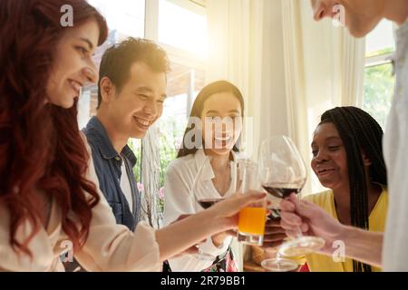 Allegri giovani amici interculturali che si sfidano con i loro drink a casa davanti alla macchina fotografica mentre si godono la festa e il divertimento Foto Stock