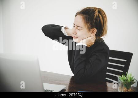 Giovane donna asiatica è seduta al lavoro e ha dolore al collo mentre è seduta in ufficio Foto Stock