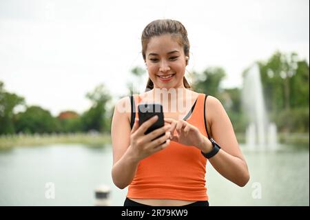 Una bella giovane donna asiatica in abbigliamento sportivo indossando auricolari Bluetooth e scegliendo una playlist musicale sul suo telefono prima di correre da sola nel parco. lei Foto Stock