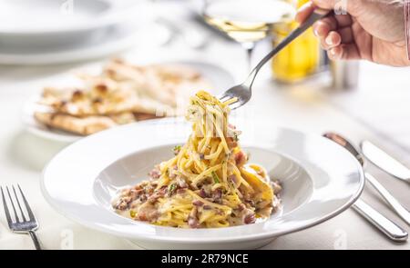 La mano di una persona prende spaghetti alla carbonara da un piatto in un ristorante. Foto Stock
