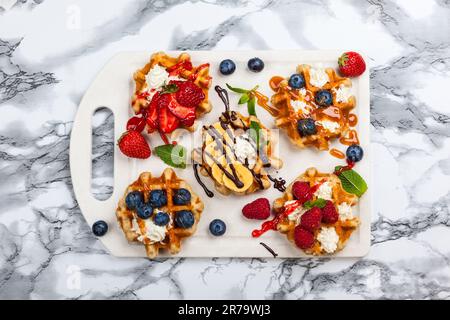 Deliziosi waffle belgi con frutti di bosco, frutta, panna montata e diversi dolcetti su marmo bianco. Vista dall'alto. Foto Stock
