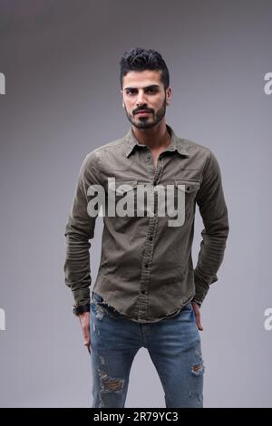Giovane in forma, attraente mediorientale, adornato da una camicia verde militare e da jeans blu perfetti. Catturato in un colpo medio, appare serio Foto Stock