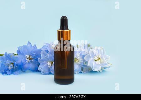 Siero in flacone di vetro marrone senza marchio con pipetta e fiore blu di delfinio su sfondo blu. Fustellato per bottiglie cosmetiche con acido ialuronico per marca Foto Stock