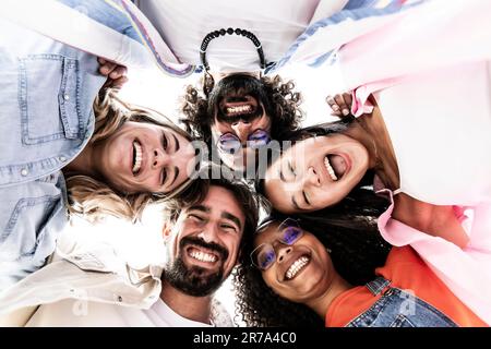 Vista bassa di un gruppo multirazziale di giovani in piedi in cerchio ridendo e fissando la macchina fotografica. Diversi Millennial felici in cerchio guardando verso il basso Foto Stock