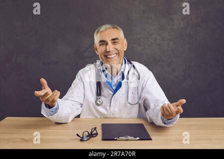 Ritratto di felice medico anziano seduto alla scrivania, guardando la macchina fotografica e sorridendo Foto Stock