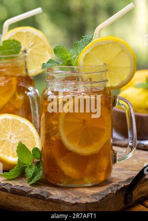 Tè ghiacciato in due vasetti di vetro con fettine di limone e menta su vassoio di legno con sfondo naturale Foto Stock
