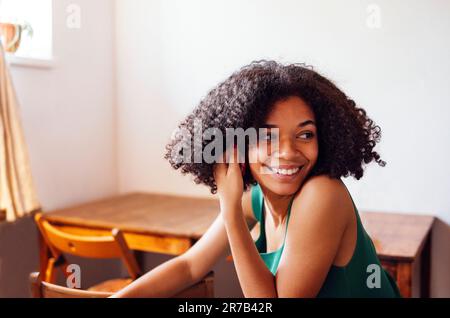 Primo piano di ritratto di carina ragazza di razza mista sorridente e tenendo la mano vicino al suo volto. Adorabile giovane africano riccio in verde canotta top interni. TA Foto Stock