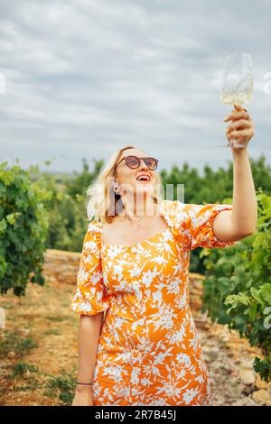 Allegra donna bionda in abito arancione brillante con stampa floreale tiene un bicchiere di champagne sopra la testa in vigna. Bella ragazza in occhiali da sole lau Foto Stock