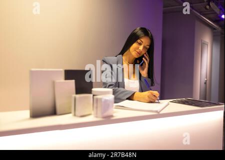 Giovane bella receptionist focalizzato parla al telefono e scrive note sul cliente. Attraente ragazza dai capelli castani si alza alla reception e fa ca Foto Stock